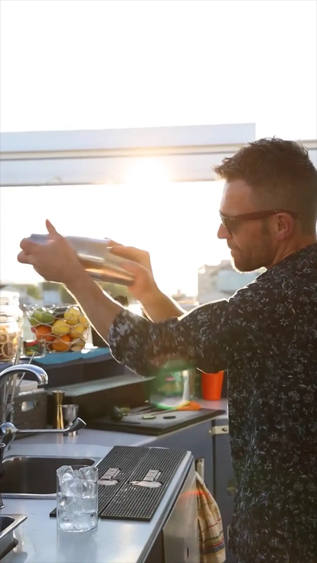 Ein Mann mit Sonnenbrille schüttelt einen Cocktailshaker in einer Küche bei Sonnenuntergang.