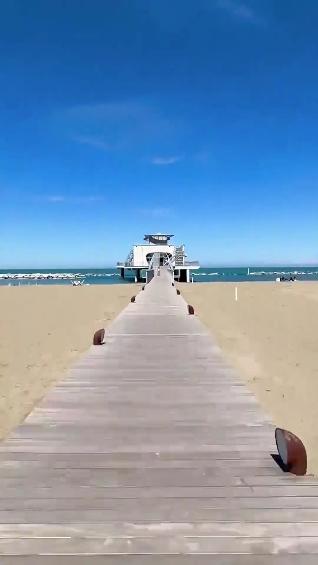 Ein hölzerner Pier führt über einen Sandstrand zu einem modernen Gebäude auf dem Meer, unter einem klaren blauen Himmel.