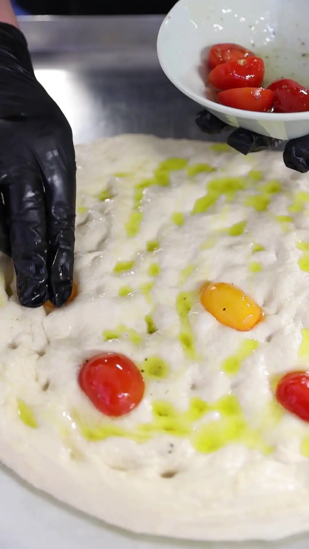 Ein Teig mit Olivenöl und Kirschtomaten wird mit Handschuhen zubereitet.